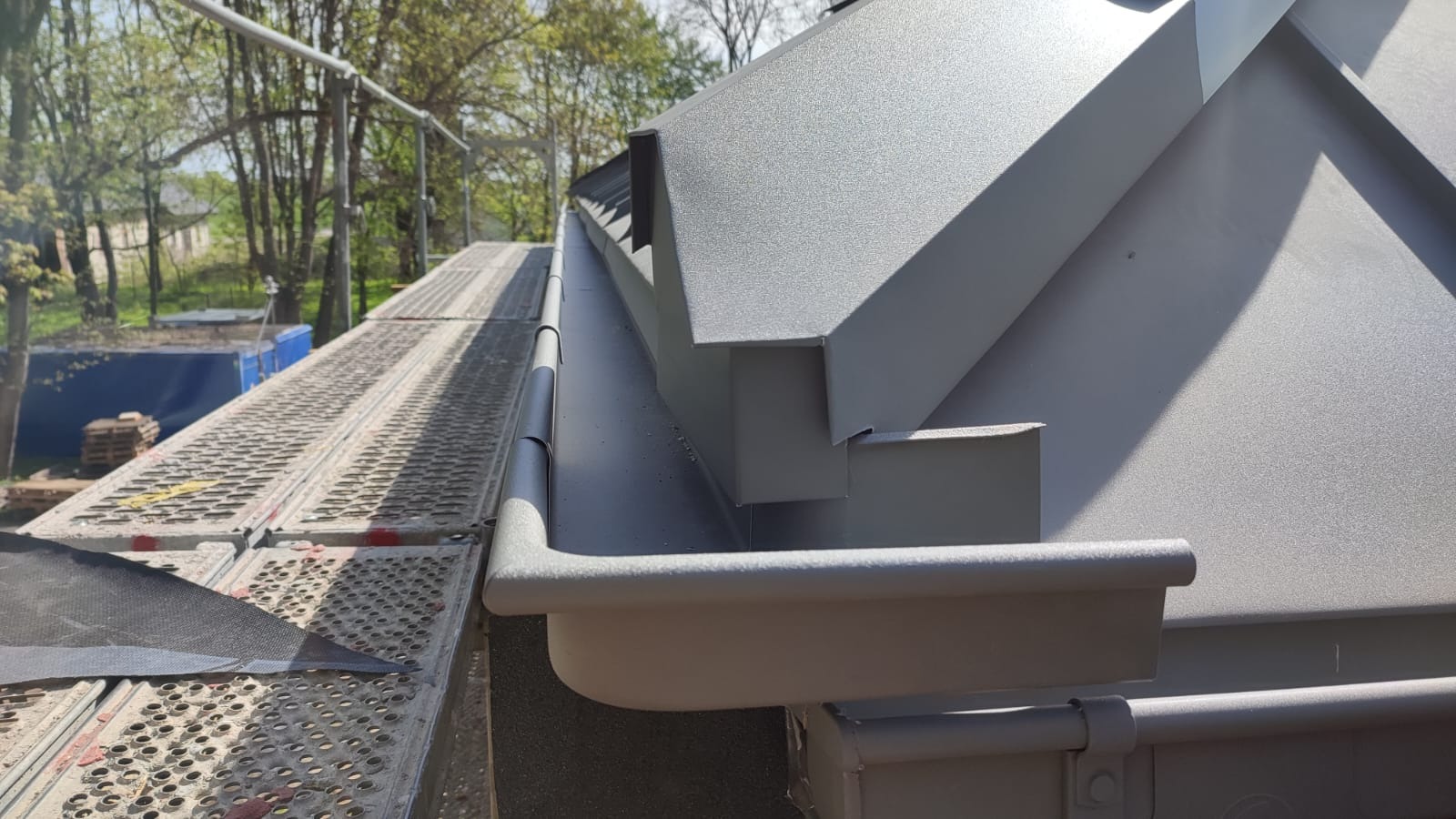 Szare, metalowe rynny zamontowane na nowym dachu, widoczne z rusztowania w słoneczny dzień.