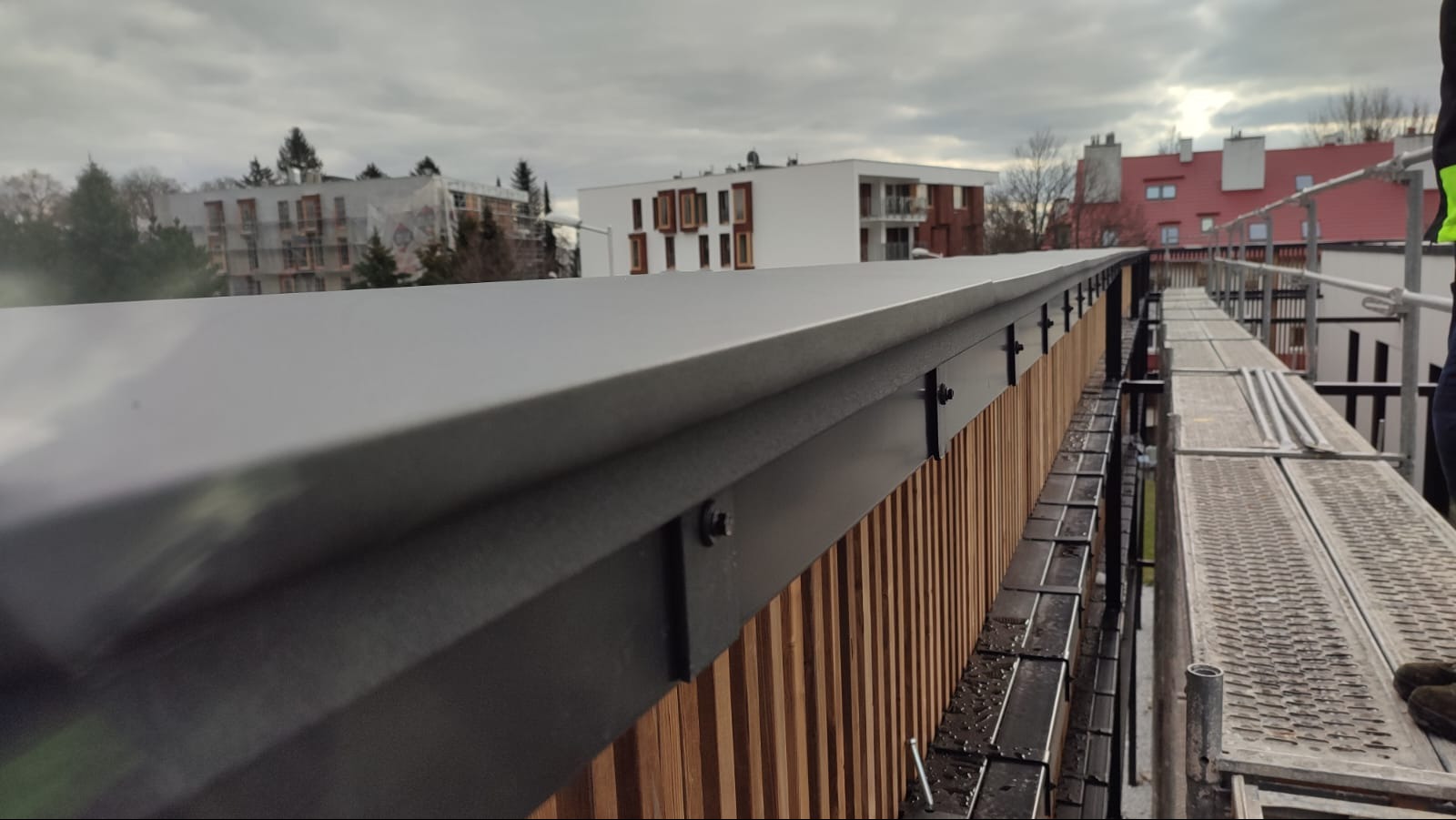 Szary, metalowy dach z drewnianą okładziną pionową, mocowany śrubami, widok z rusztowania, w tle budynki mieszkalne pod pochmurnym niebem.