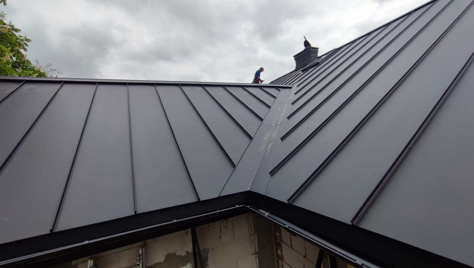 Nowoczesne, grafitowe pokrycie dachowe z blachy na budynku w trakcie budowy, widoczny komin i pracownik na dachu.