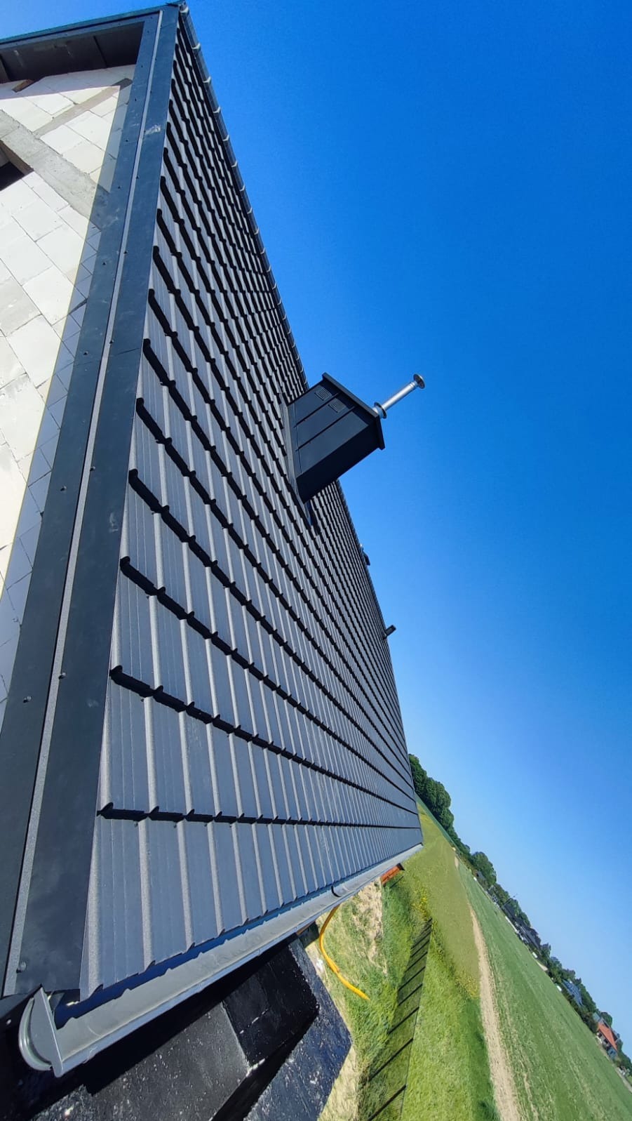 Nowy, grafitowy dach z blachodachówki modułowej, z kominkiem wentylacyjnym, na tle błękitnego nieba i zielonego pola, widok z dołu.