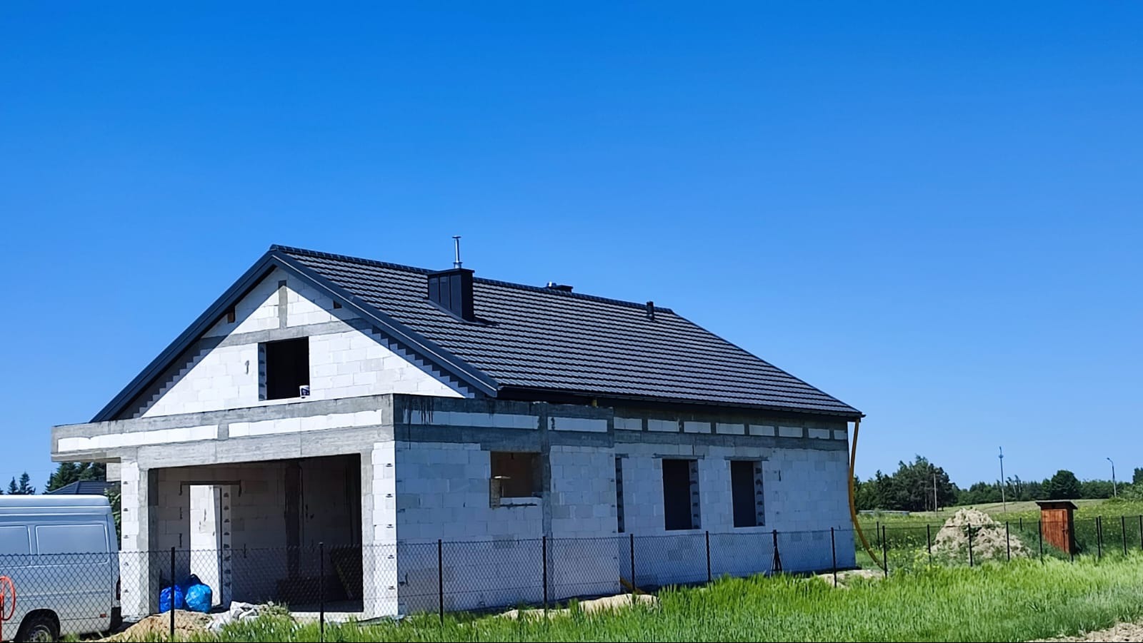 Nowy dom w trakcie budowy, z ciemnoszarą dachówką i kominem, widoczne otwory okienne i drzwiowe, ogrodzony siatką, z niebieskimi workami w garażu i dostawczym samochodem w tle.