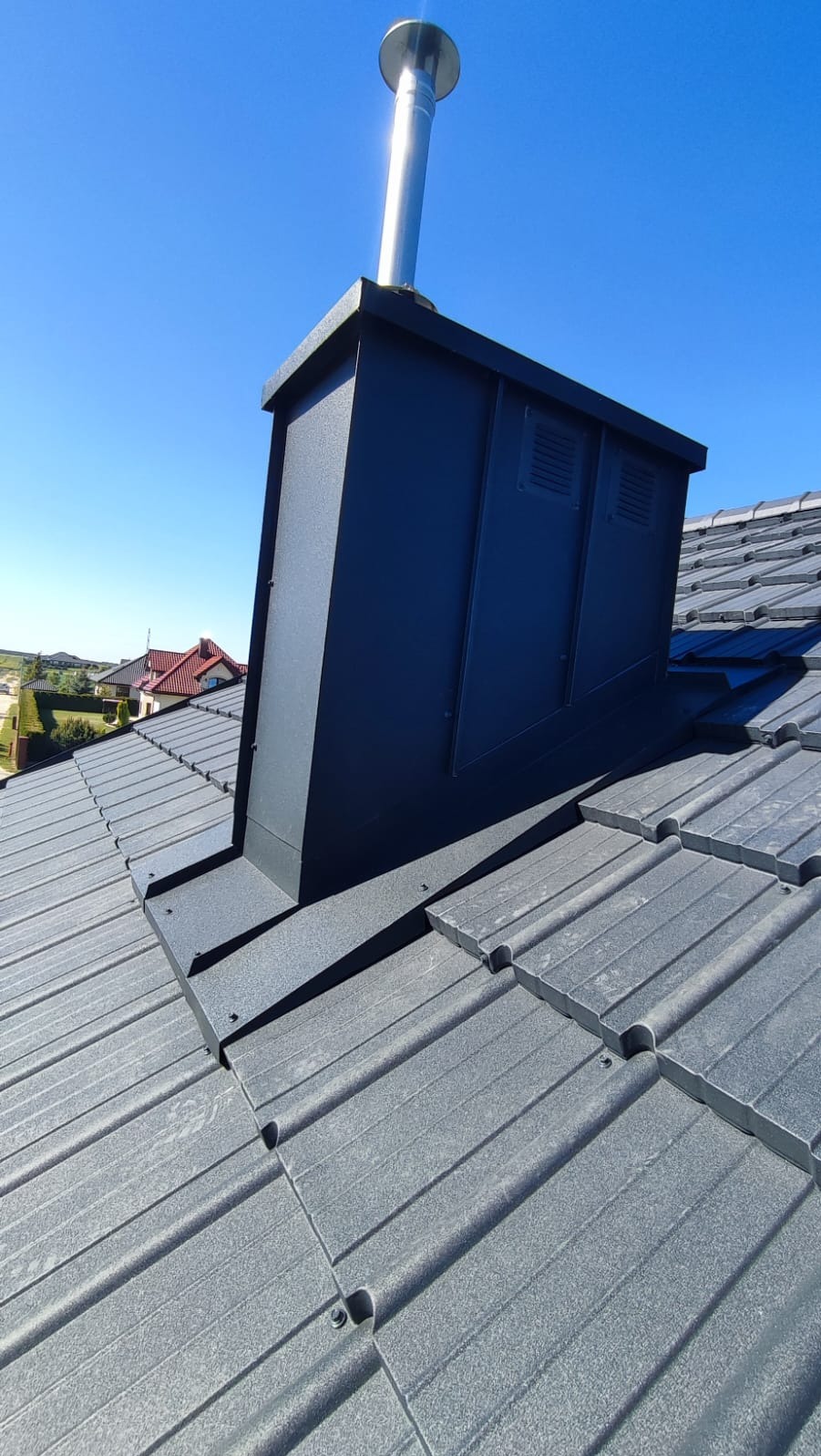 Szary dach z metalową obudową komina wentylacyjnego i rurą w słoneczny dzień.