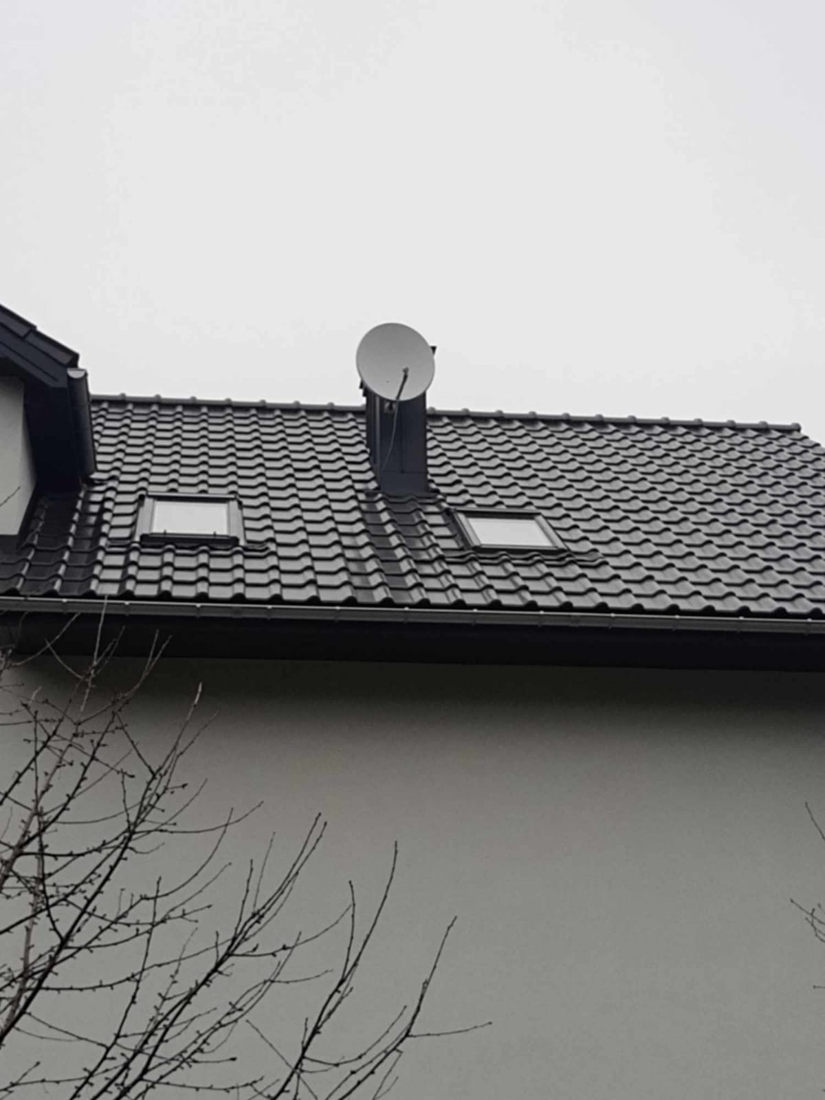 Czarny dach z dachówki ceramicznej z dwoma oknami dachowymi i anteną satelitarną zamontowaną na kominie, widok z dołu na tle szarego nieba.