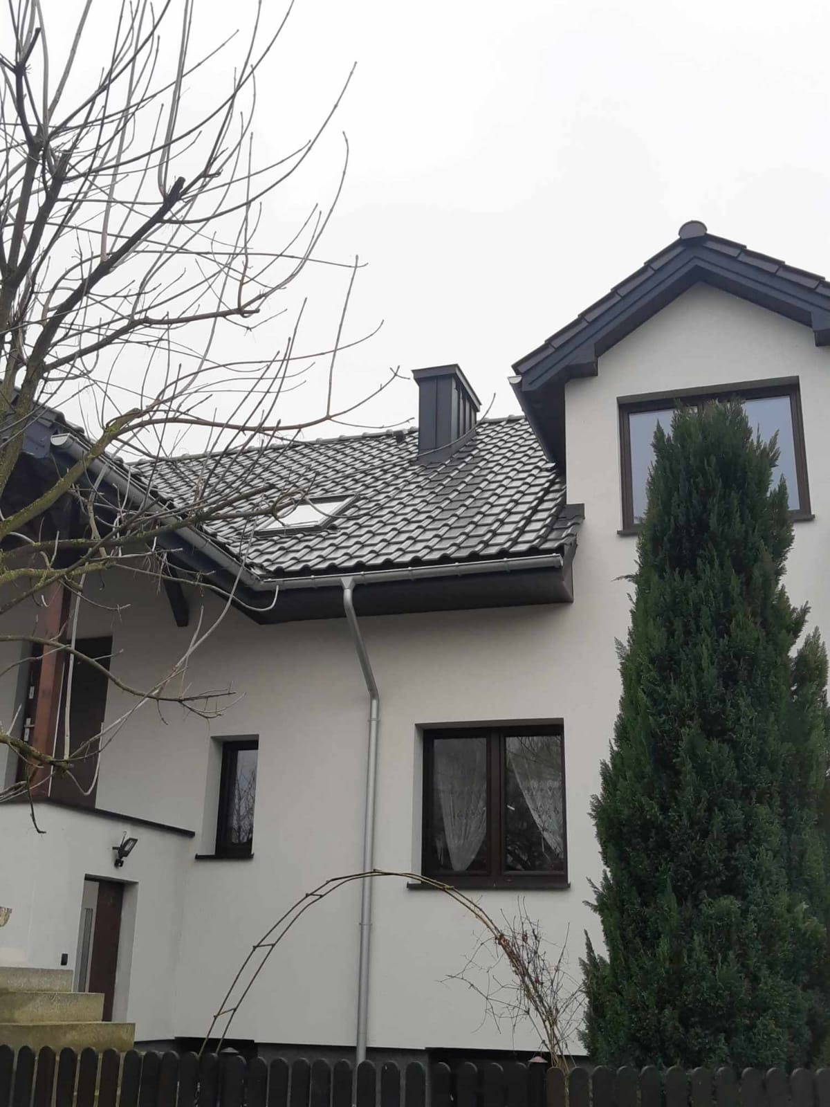 Dwukondygnacyjny dom z ciemnoszarą dachówką ceramiczną, widoczne okno dachowe, rynna, komin, oraz drzewa iglaste i liściaste bez liści. Biała elewacja i ciemne obramowania okien.