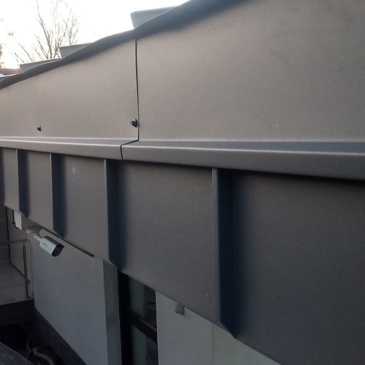 Szare, metalowe obróbki blacharskie dachu z widocznymi śrubami mocującymi i fragmentem kamery monitoringu na ścianie budynku.