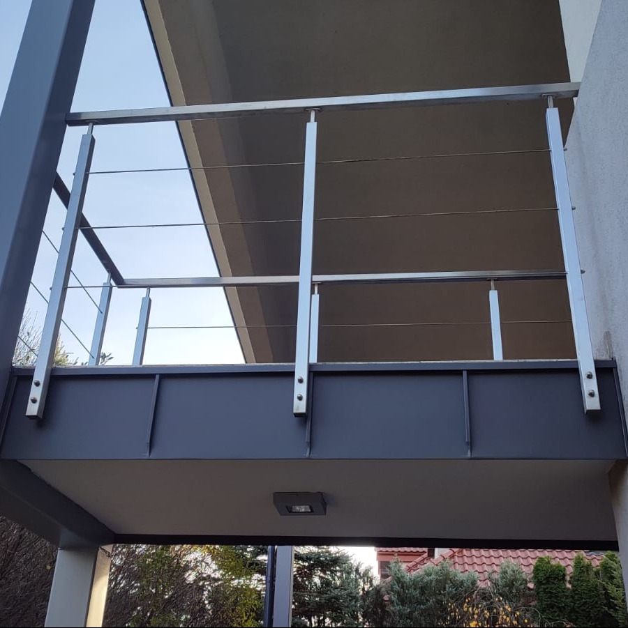 Nowoczesna balustrada ze stali nierdzewnej z poziomymi linkami, zamontowana na ciemnoszarej konstrukcji balkonu, widok z dołu na tle zieleni.