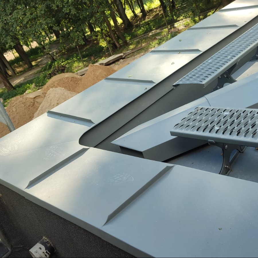 Szary dach z blachy na tle zieleni drzew, widoczne obróbki blacharskie i ślady butów na powierzchni.