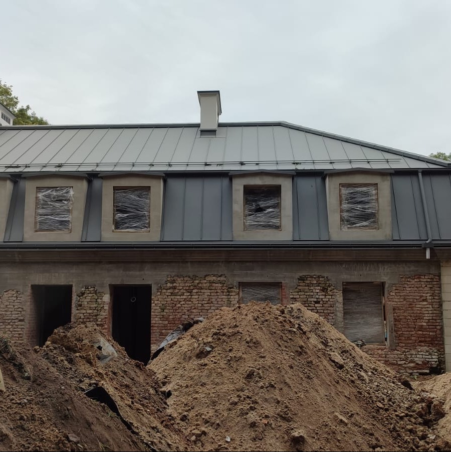 Budynek w trakcie remontu dachu z nowym, szarym pokryciem blacharskim i zabezpieczonymi oknami na poddaszu; widoczne ceglane ściany i hałda ziemi przed budynkiem.