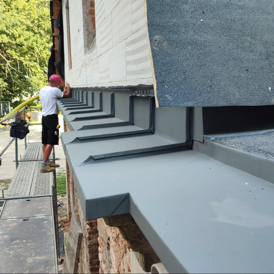 Pracownik na rusztowaniu montuje szare, metalowe obróbki blacharskie na krawędzi dachu budynku z ceglaną ścianą, widoczne elementy dachu z papy.