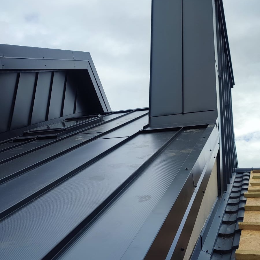 Nowoczesny, grafitowy dach z blachy na rąbek stojący, z oknem dachowym i obróbką komina, widok z dołu na tle pochmurnego nieba.