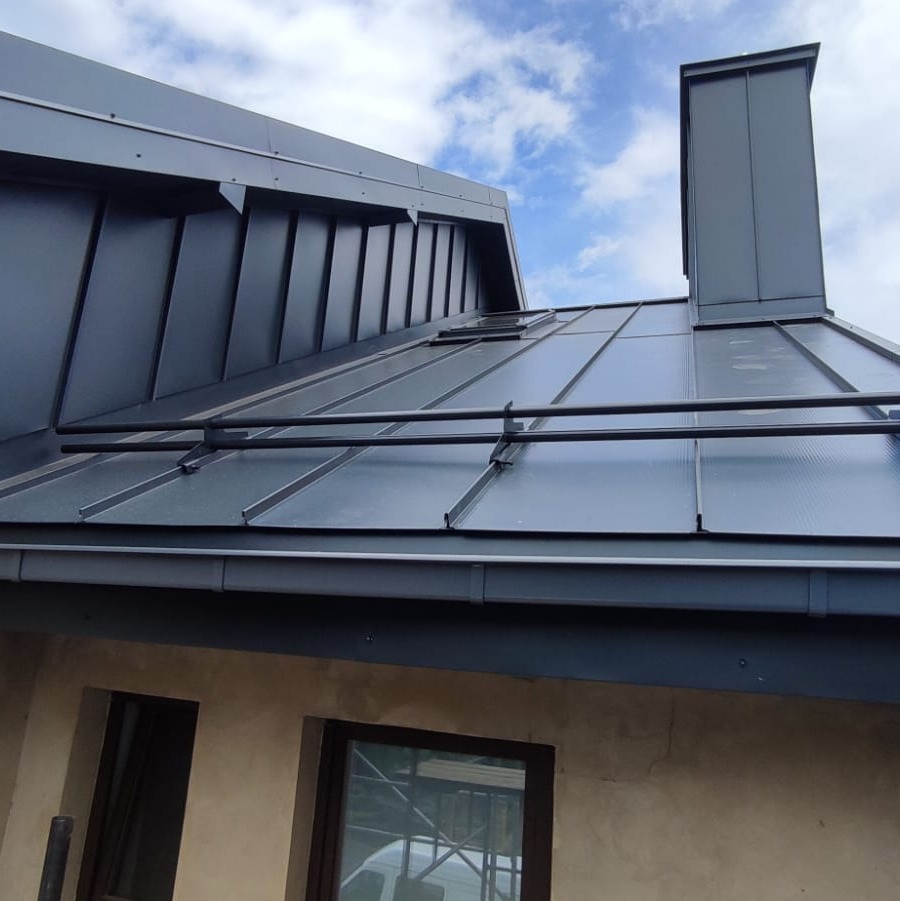 Nowoczesny dach z blachy na rąbek stojący, system click Ruukki, z widocznym kominem i systemem zabezpieczeń przeciwśniegowych, na tle błękitnego nieba.