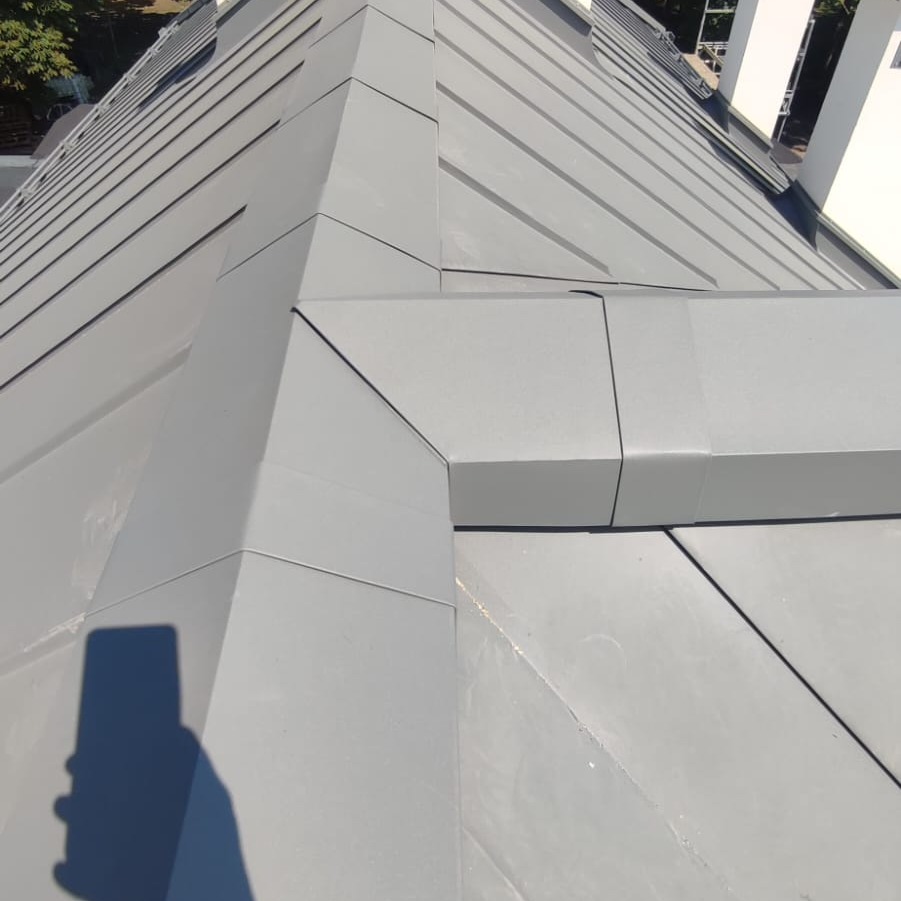 Szary dach z blachy trapezowej z obróbkami blacharskimi przy kominie, widoczny cień fotografa.