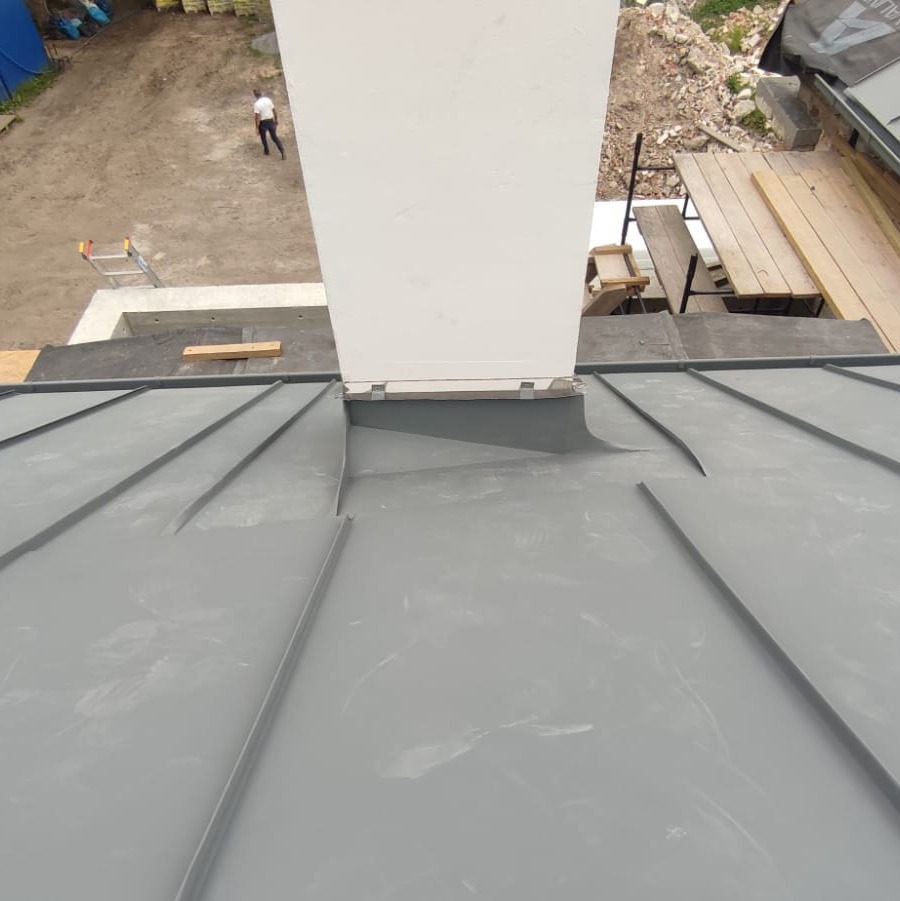 Szare metalowe poszycie dachu z pionowym kominem w trakcie montażu, widok z góry na fragment dachu i otoczenie budowy.