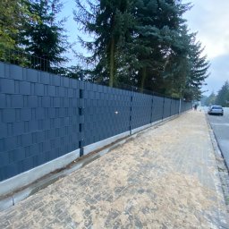 Pan Płot Kamil Kot - Długi, grafitowy płot panelowy z metalową siatką u góry, wzdłuż chodnika z kostki brukowej, z widocznymi drzewami w tle i osobą z psem na ulicy.