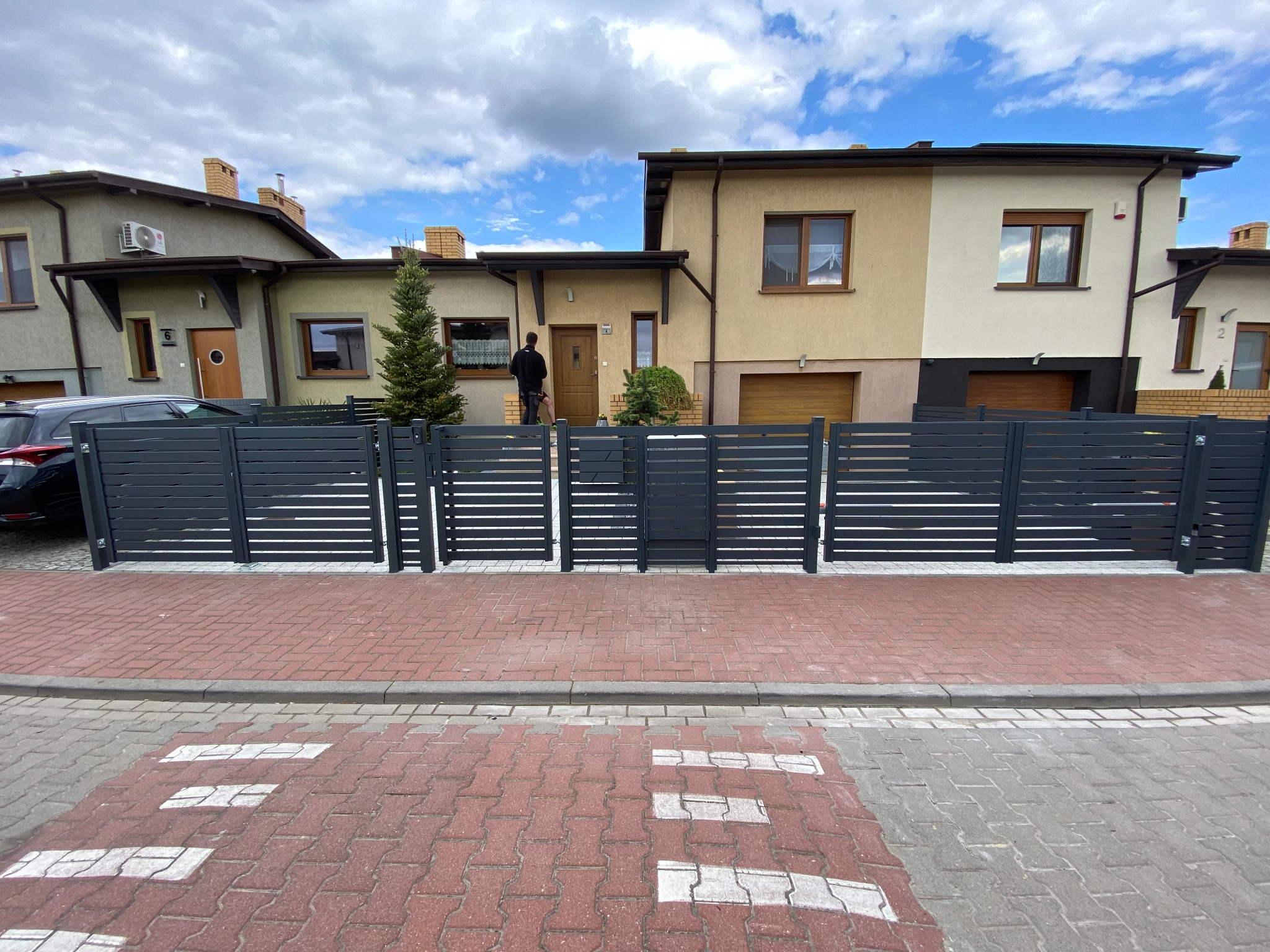 Nowoczesne ogrodzenie panelowe w kolorze grafitowym, z poziomymi szczeblami, przed domami jednorodzinnymi, widok z poziomu ulicy z kostki brukowej.