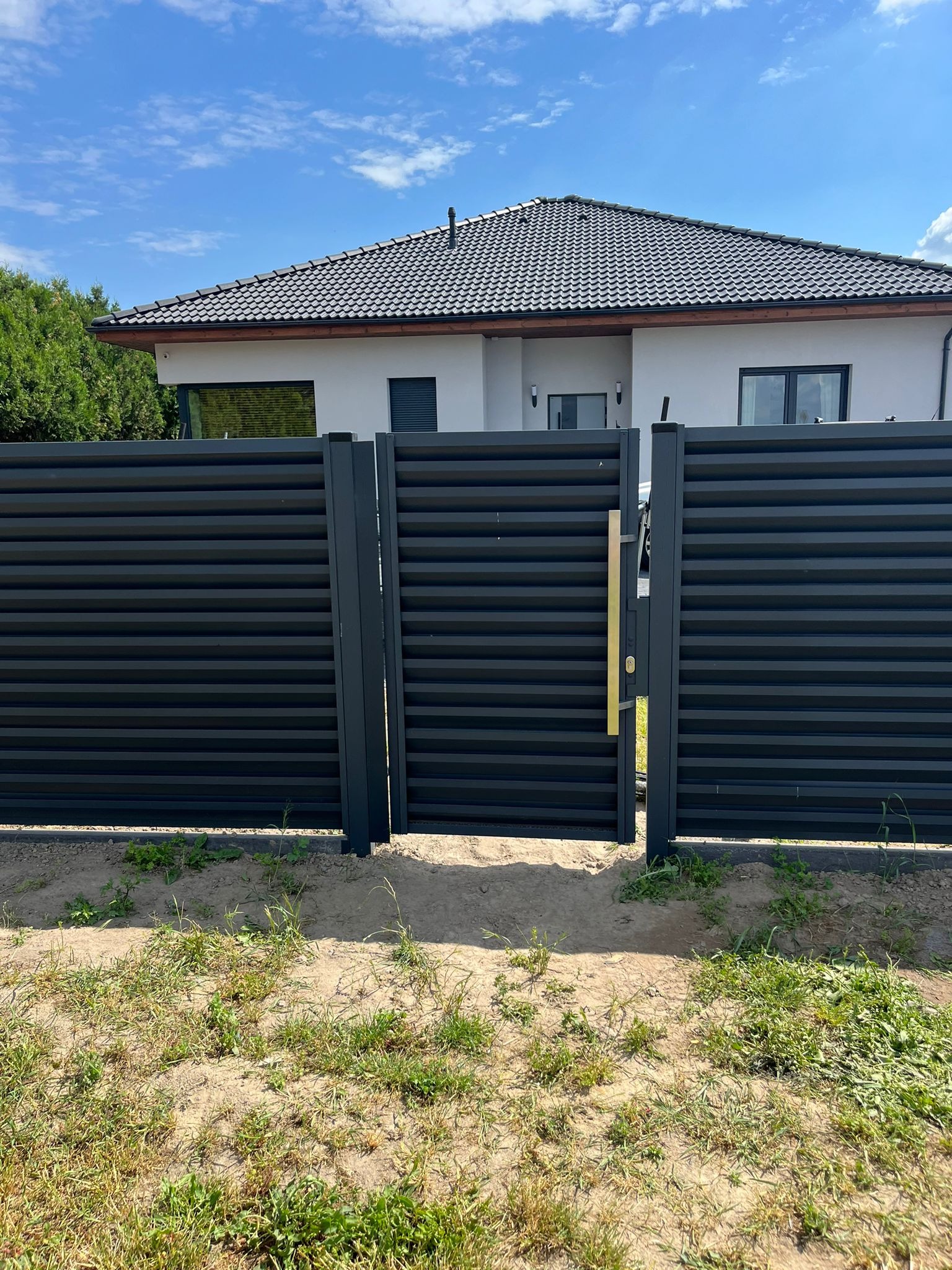 Nowoczesna, pozioma, czarna brama metalowa z złotą klamką, prowadząca na posesję z domem o szarej dachówce.