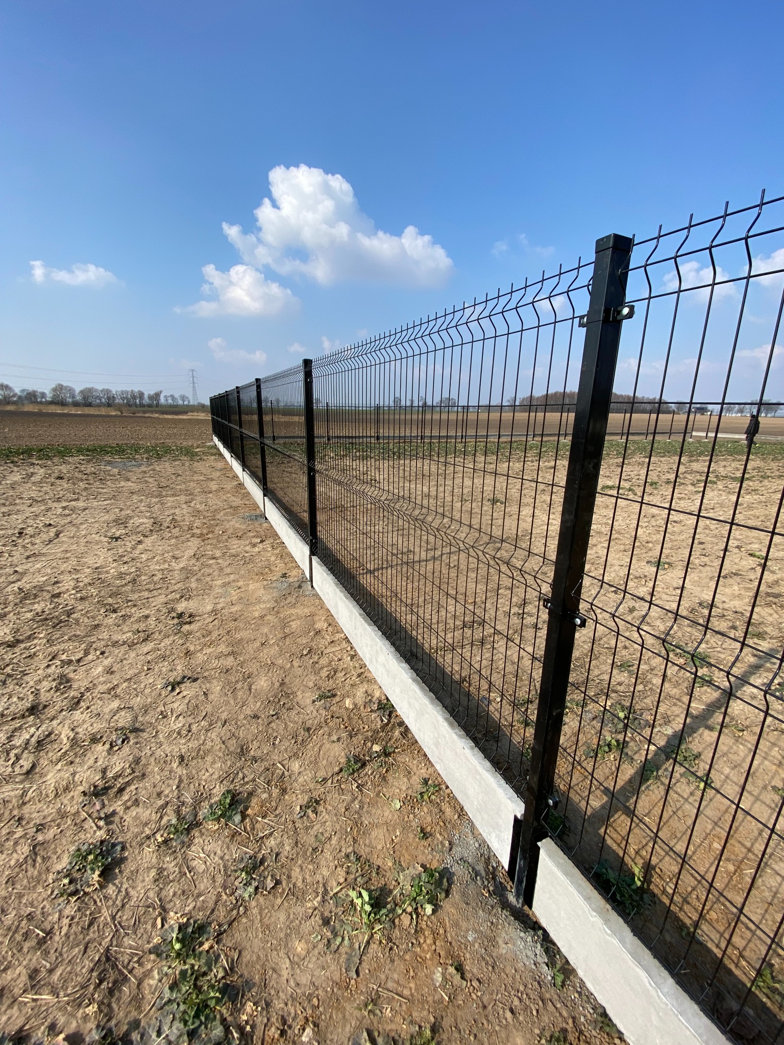 Długa linia czarnego ogrodzenia panelowego z betonową podmurówką na tle pola i błękitnego nieba z chmurami.