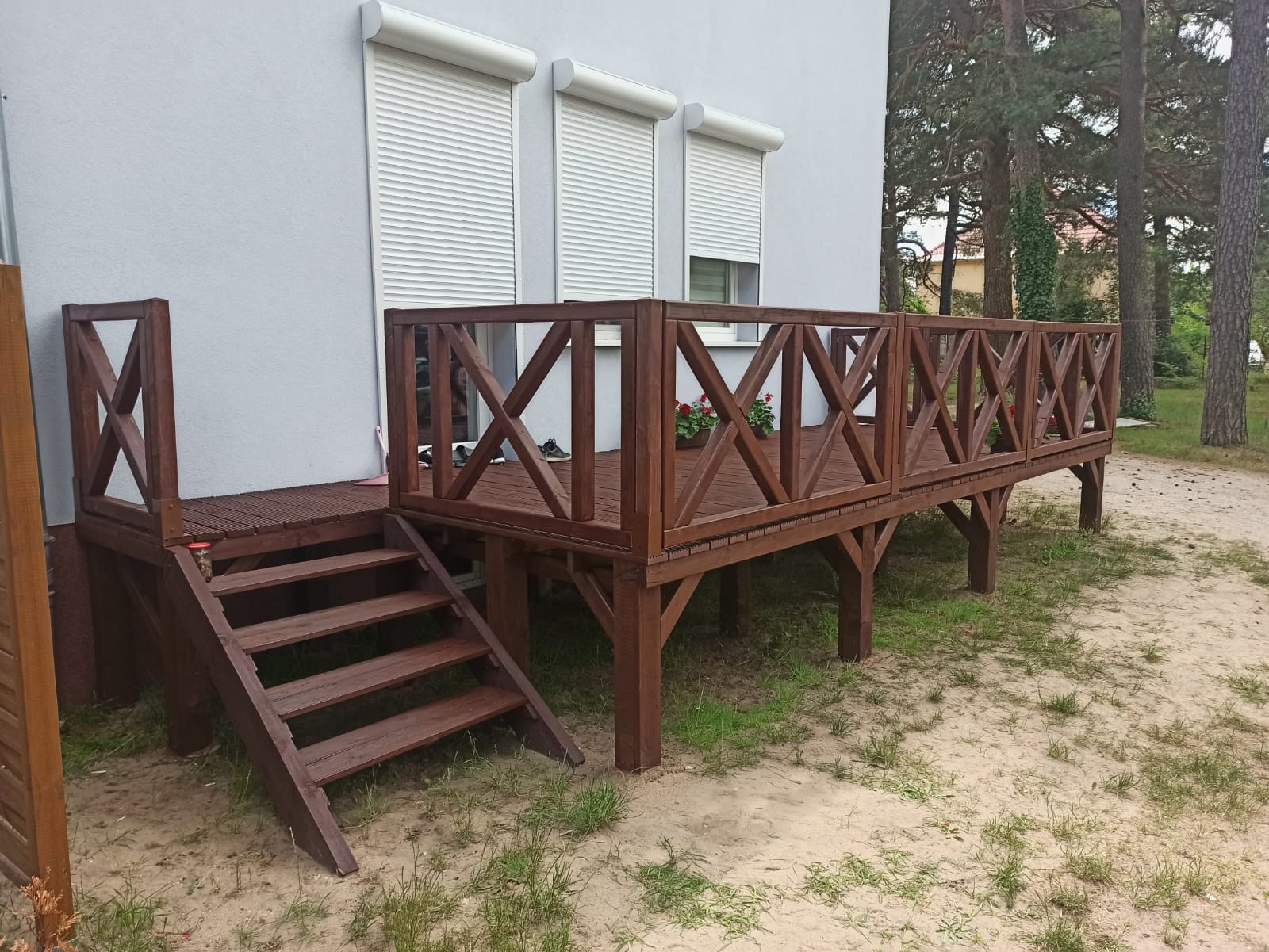 Drewniany taras z balustradą w kratkę, schodami i doniczkami z kwiatami, przylegający do jasnego budynku z roletami zewnętrznymi, otoczony piaszczystym terenem i drzewami.