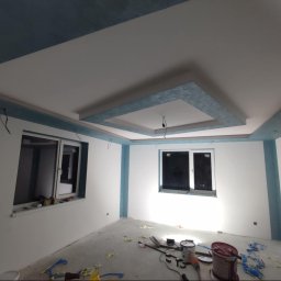 WRO IVESTALL - Pokój po częściowym odświeżeniu: białe ściany z błękitnymi akcentami na suficie i wokół okien, widoczne narzędzia malarskie na podłodze, sufit z dekoracyjnym podwieszanym elementem w kolorze...