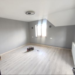 WRO IVESTALL - Szare wnętrze pokoju po malowaniu ścian i położeniu jasnej podłogi laminowanej, widoczny grzejnik i okno w skosie dachu, na podłodze stoi boombox.