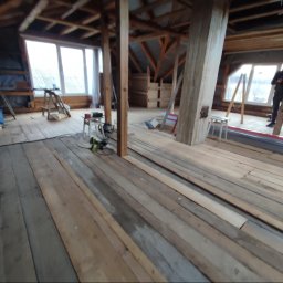 WRO IVESTALL - Poddasze w trakcie remontu, widoczne drewniane belki konstrukcyjne, częściowo ułożona podłoga z desek, narzędzia i materiały budowlane, osoba w tle.