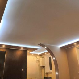 WRO IVESTALL - Nowoczesne wnętrze z podwieszanym sufitem, oświetleniem LED i łukowym przejściem między pomieszczeniami. Widoczny fragment ściany z ciemną tapetą i jasne ściany w drugim pomieszczeniu, gdzie...