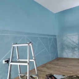 Niebieska ściana z geometrycznym wzorem, wykonana techniką dekoracyjną, widoczna aluminiowa drabina i fragment podłogi z paneli.