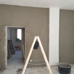 WRO IVESTALL - Ściana w trakcie nakładania gładzi, widoczny fragment drabiny z zawieszonym wiadrem z zaprawą, oraz wiadro stojące na podłodze. W tle widoczne materiały budowlane i otwór drzwiowy.