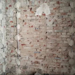 Surowa ściana z cegły, przygotowana pod montaż płyt kartonowo-gipsowych, z widocznymi metalowymi profilami i śladami zaprawy.