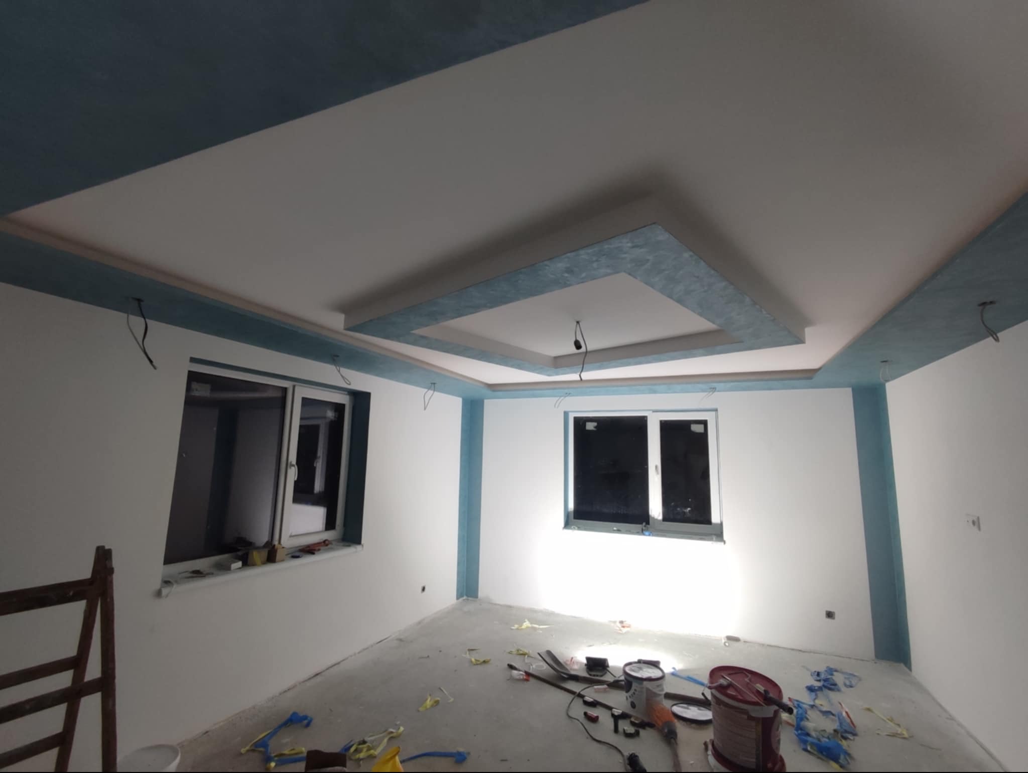 Pokój po częściowym odświeżeniu: białe ściany z błękitnymi akcentami na suficie i wokół okien, widoczne narzędzia malarskie na podłodze, sufit z dekoracyjnym podwieszanym elementem w kolorze...