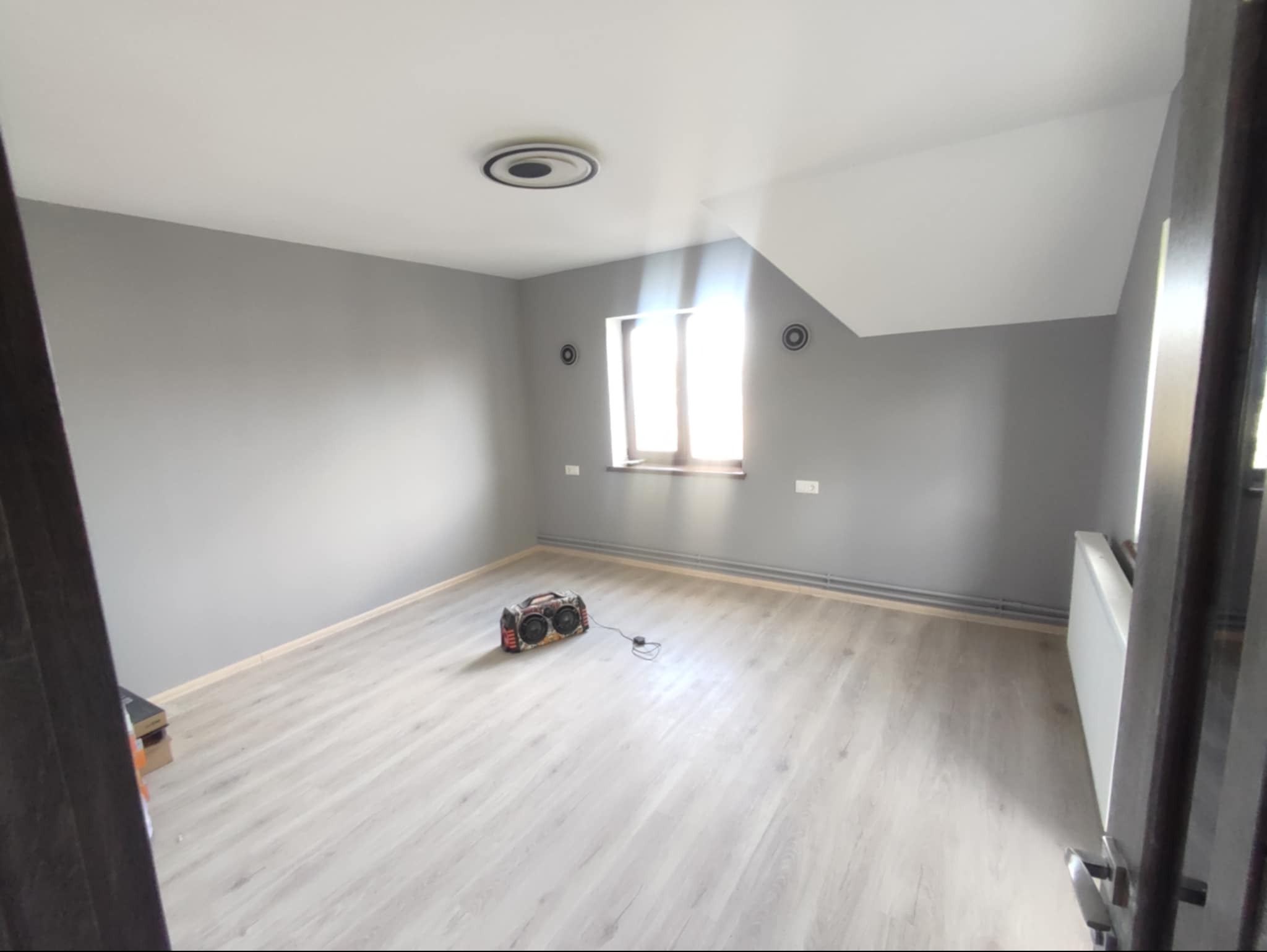 Szare wnętrze pokoju po malowaniu ścian i położeniu jasnej podłogi laminowanej, widoczny grzejnik i okno w skosie dachu, na podłodze stoi boombox.