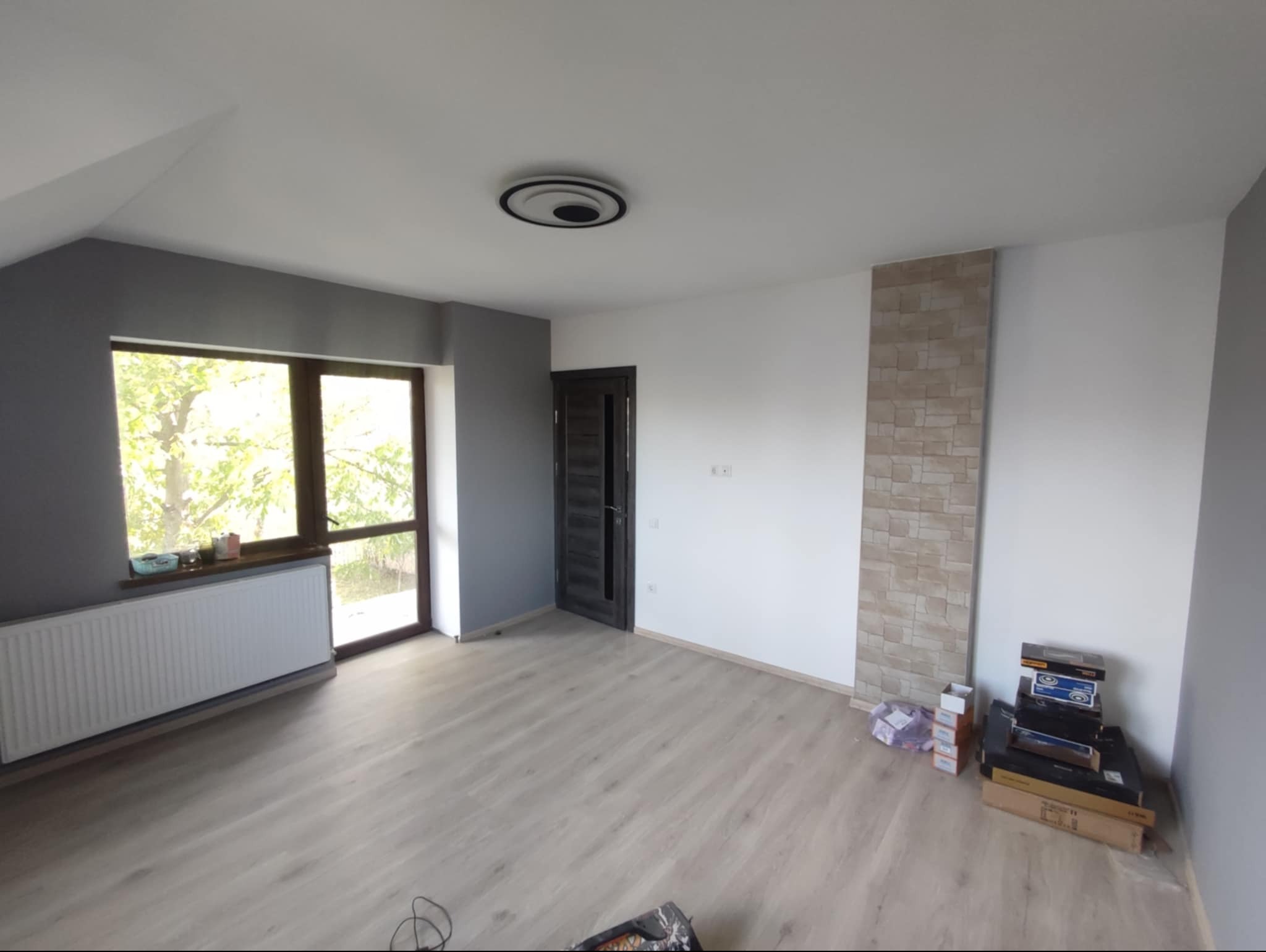 Wnętrze pokoju po remoncie: szare ściany, biała ściana z dekoracyjnym panelem imitującym cegłę, jasna podłoga, grzejnik pod oknem, drzwi wejściowe, lampa sufitowa i kartony z materiałami...