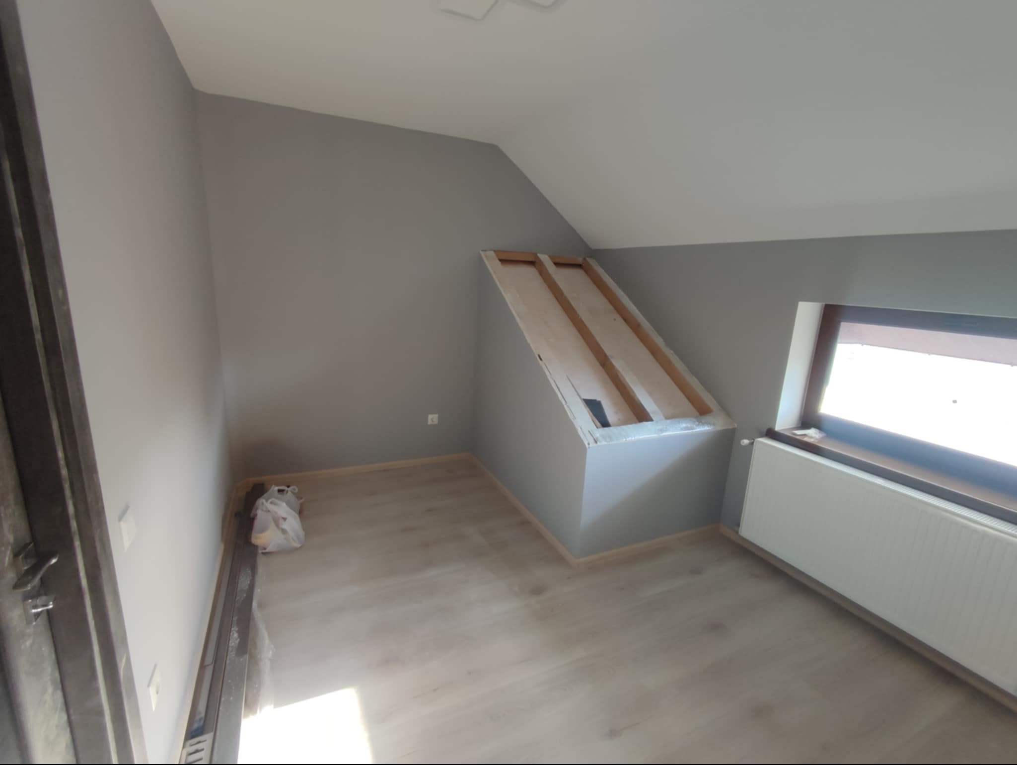 Pomalowane na szaro poddasze ze skosami, widoczne elementy konstrukcji zabudowy wnęki, grzejnik pod oknem i torba z materiałami budowlanymi w rogu.