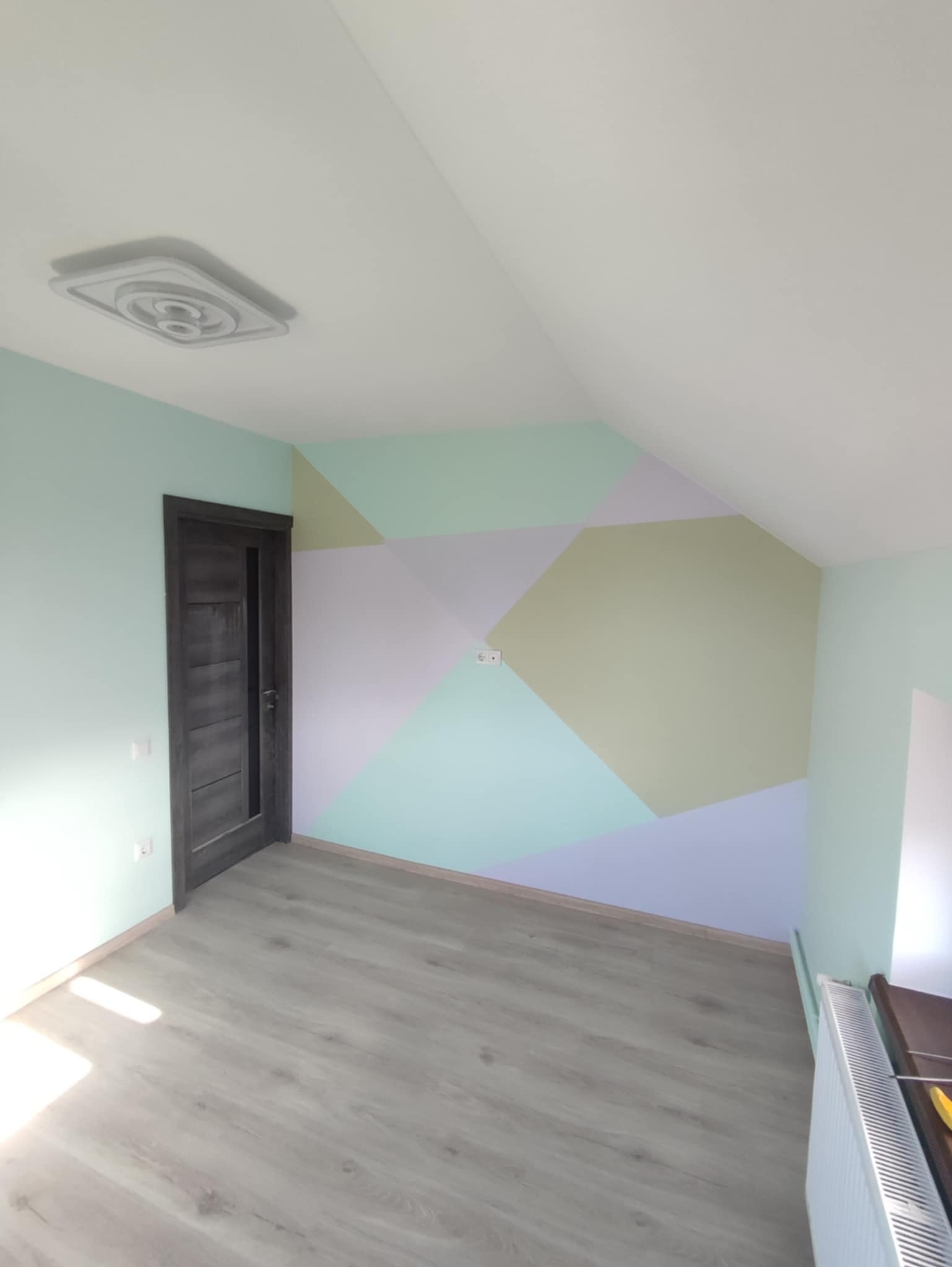Pokój na poddaszu po odświeżeniu: ściana z geometrycznym wzorem w pastelowych odcieniach zieleni, fioletu i żółci, szara drewniana podłoga, ciemne drzwi z przeszkleniami i biały grzejnik.