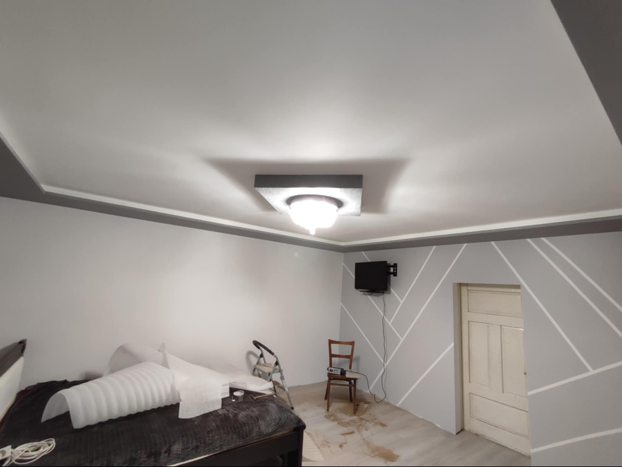 Pokój po odświeżeniu: białe ściany z szarymi geometrycznymi wzorami, szary sufit z dekoracyjnym oświetleniem, widoczne ślady po remoncie na podłodze, krzesło i drabina w tle.
