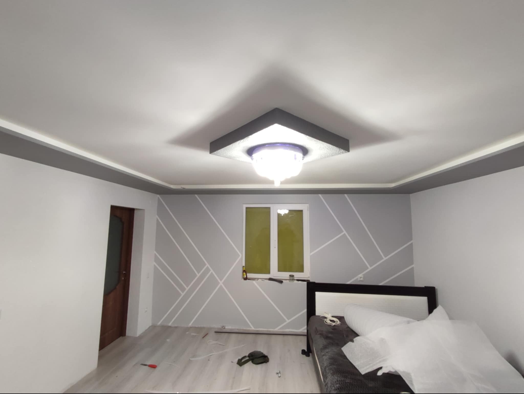 Pomieszczenie po odświeżeniu: biały sufit z szarą listwą ozdobną, nowoczesna lampa sufitowa, szare ściany z geometrycznym wzorem z białych linii. Widoczny fragment okna i łóżka z zabezpieczeniem...
