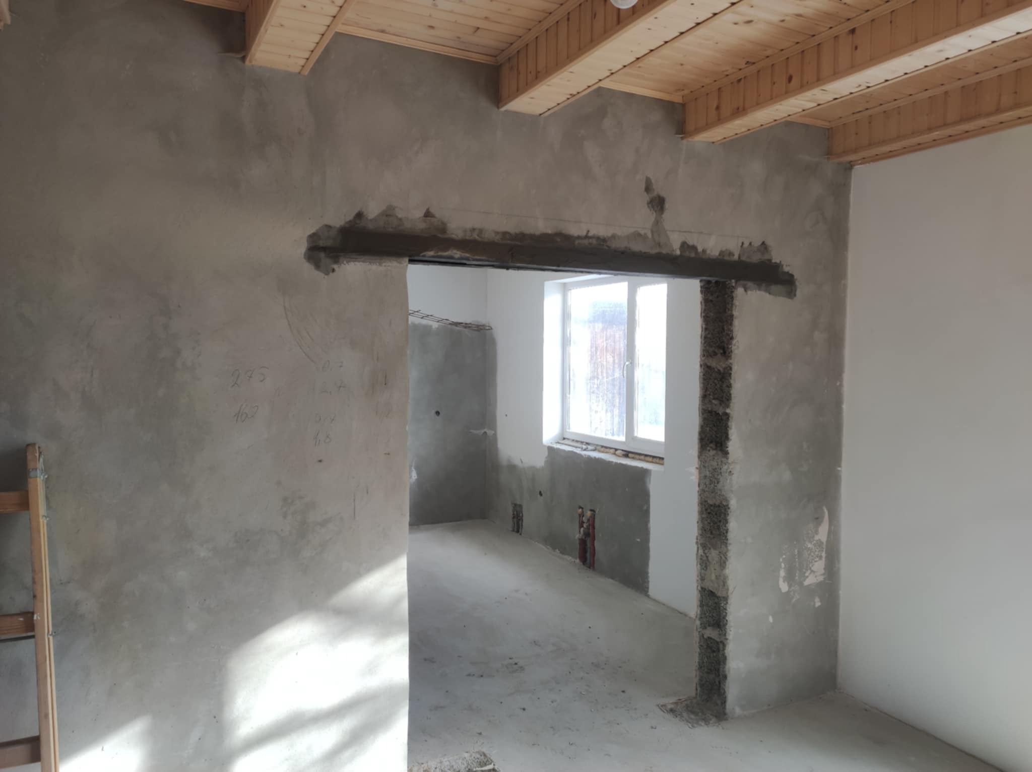 Surowy stan wnętrza w trakcie prac: ściany pokryte szarym tynkiem, drewniany sufit, przygotowane nadproże drzwiowe z widoczną strukturą bloczków, częściowo otynkowane, zarys instalacji elektrycznej...