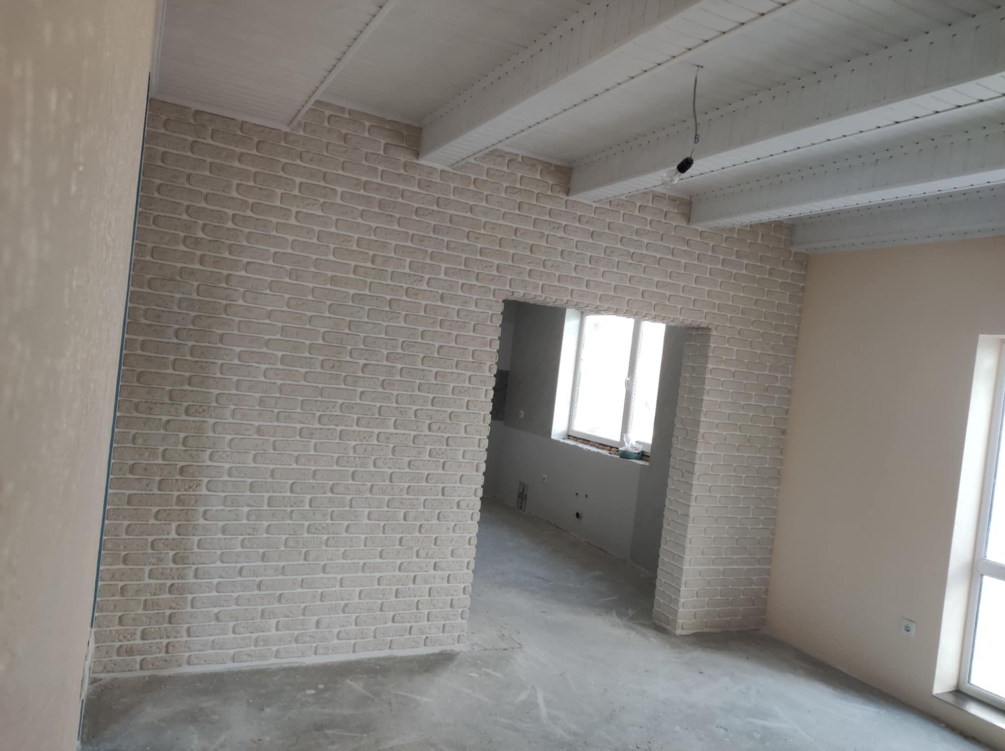 Wykończone wnętrze z dekoracyjną ścianą z cegły w odcieniach beżu i białym drewnianym sufitem, widoczne okno i surowa podłoga.