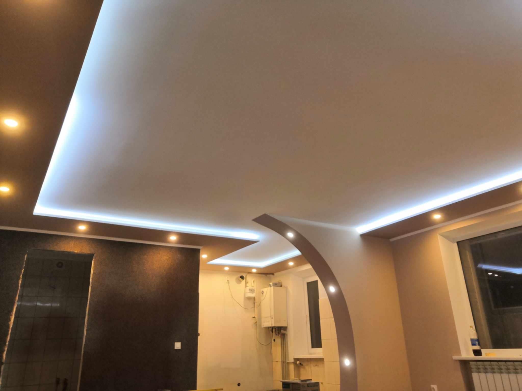 Nowoczesne wnętrze z podwieszanym sufitem, oświetleniem LED i łukowym przejściem między pomieszczeniami. Widoczny fragment ściany z ciemną tapetą i jasne ściany w drugim pomieszczeniu, gdzie...