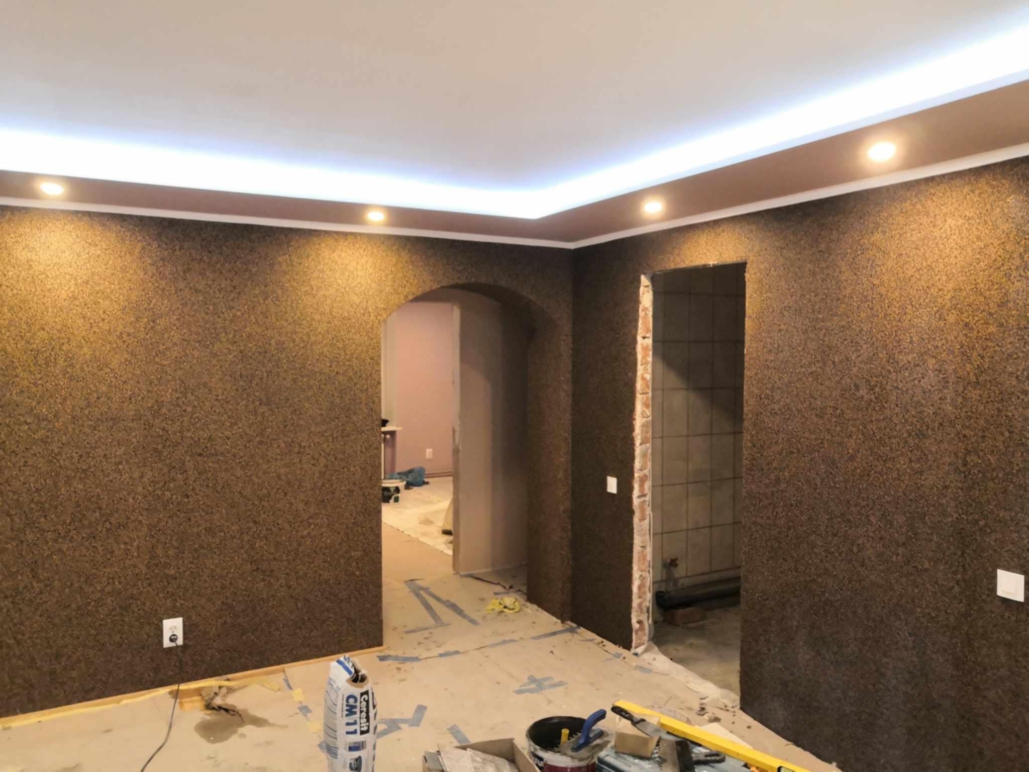 Pokój w trakcie wykańczania z brązowymi ścianami o strukturze korka, podwieszanym sufitem z oświetleniem LED, widoczne materiały budowlane i narzędzia na podłodze, oraz przejścia do innych...