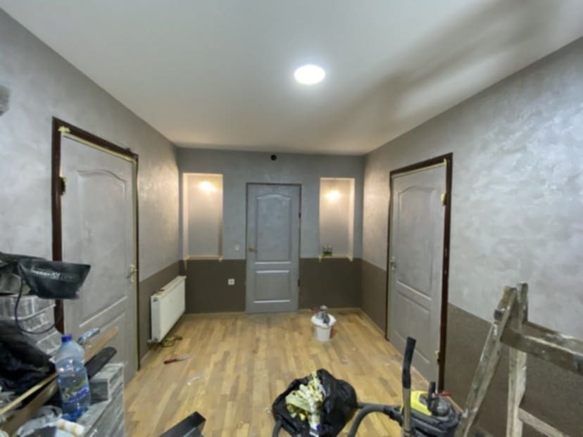 Korytarz w trakcie prac wykończeniowych: ściany częściowo pomalowane na szaro z efektem dekoracyjnym, dolna część w kolorze brązowym, widoczne drzwi z obramowaniami, grzejnik i narzędzia malarskie...