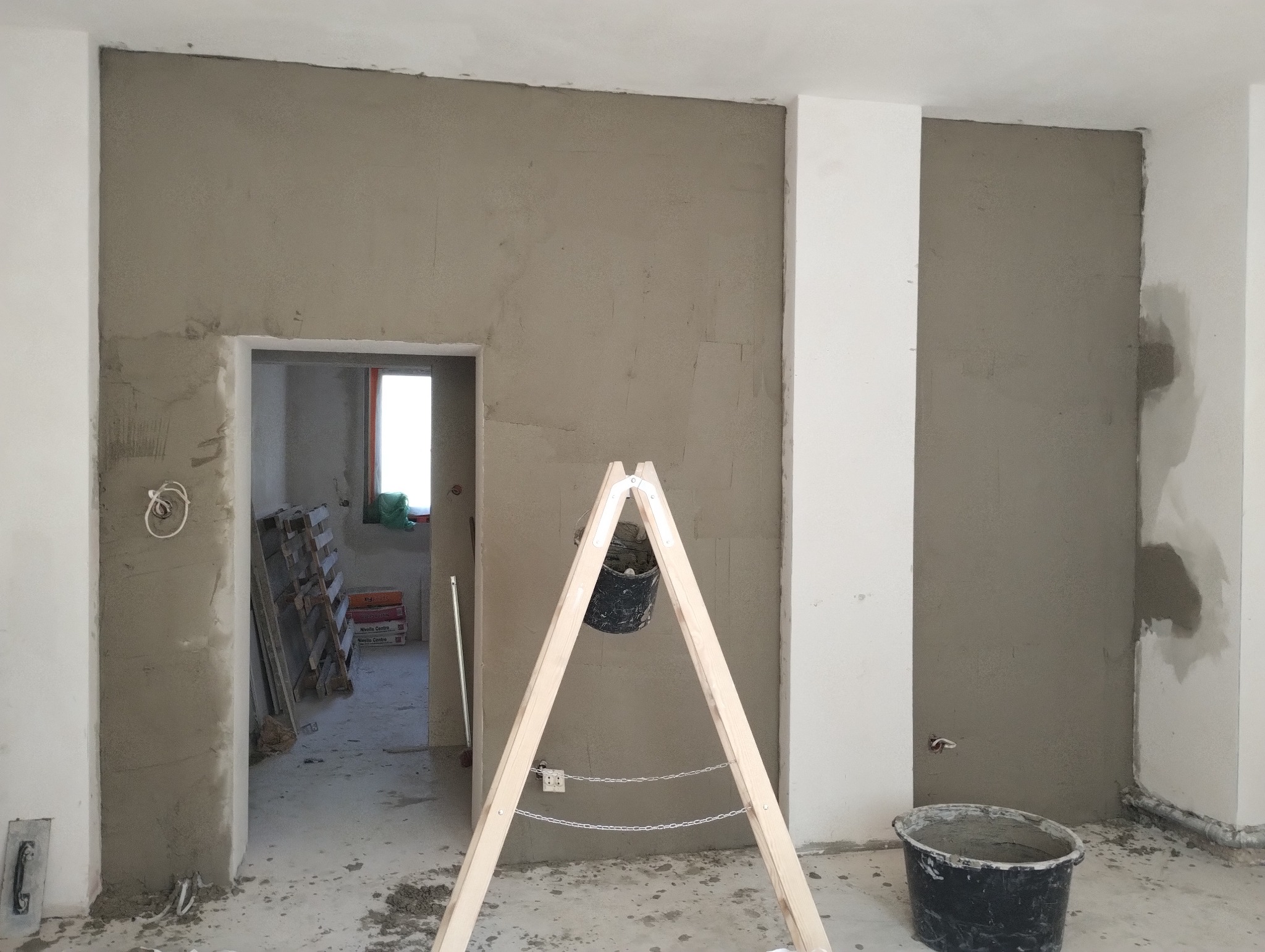 Ściana w trakcie nakładania gładzi, widoczny fragment drabiny z zawieszonym wiadrem z zaprawą, oraz wiadro stojące na podłodze. W tle widoczne materiały budowlane i otwór drzwiowy.