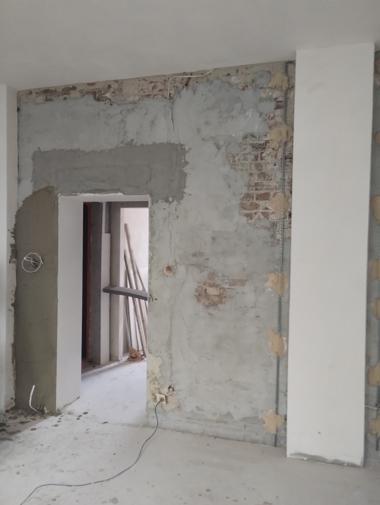 Ściana z fragmentami odsłoniętej cegły i nałożonym tynkiem, widoczny otwór drzwiowy w trakcie prac wykończeniowych, wystające kable elektryczne, surowe wnętrze.