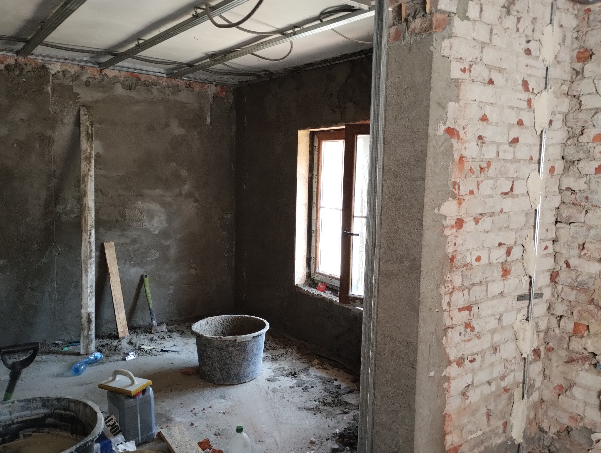 Wnętrze w trakcie remontu: otynkowana ściana, fragment odsłoniętej cegły, metalowe profile sufitowe i kubeł z zaprawą na podłodze.