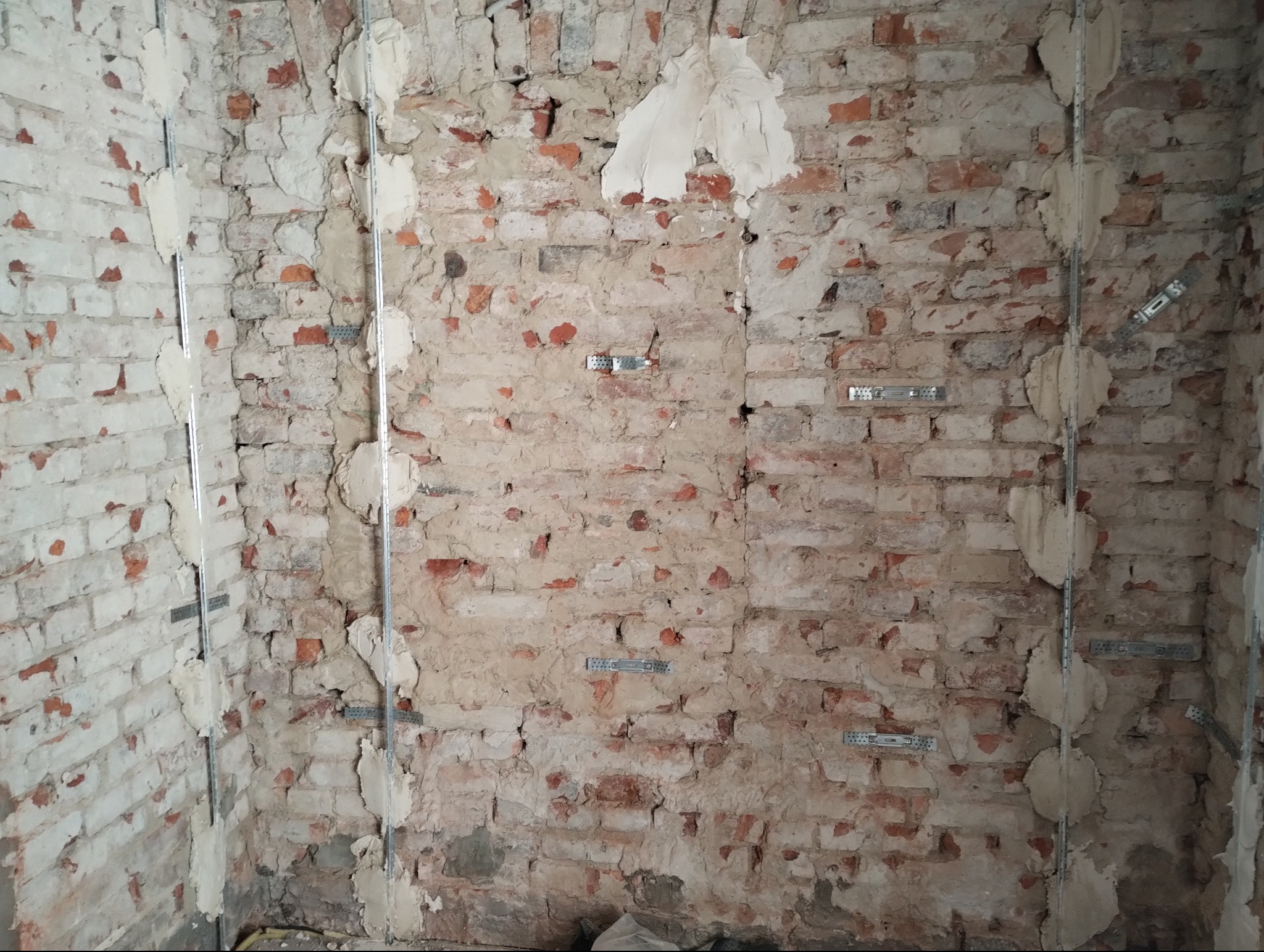 Surowa ściana z cegły, przygotowana pod montaż płyt kartonowo-gipsowych, z widocznymi metalowymi profilami i śladami zaprawy.