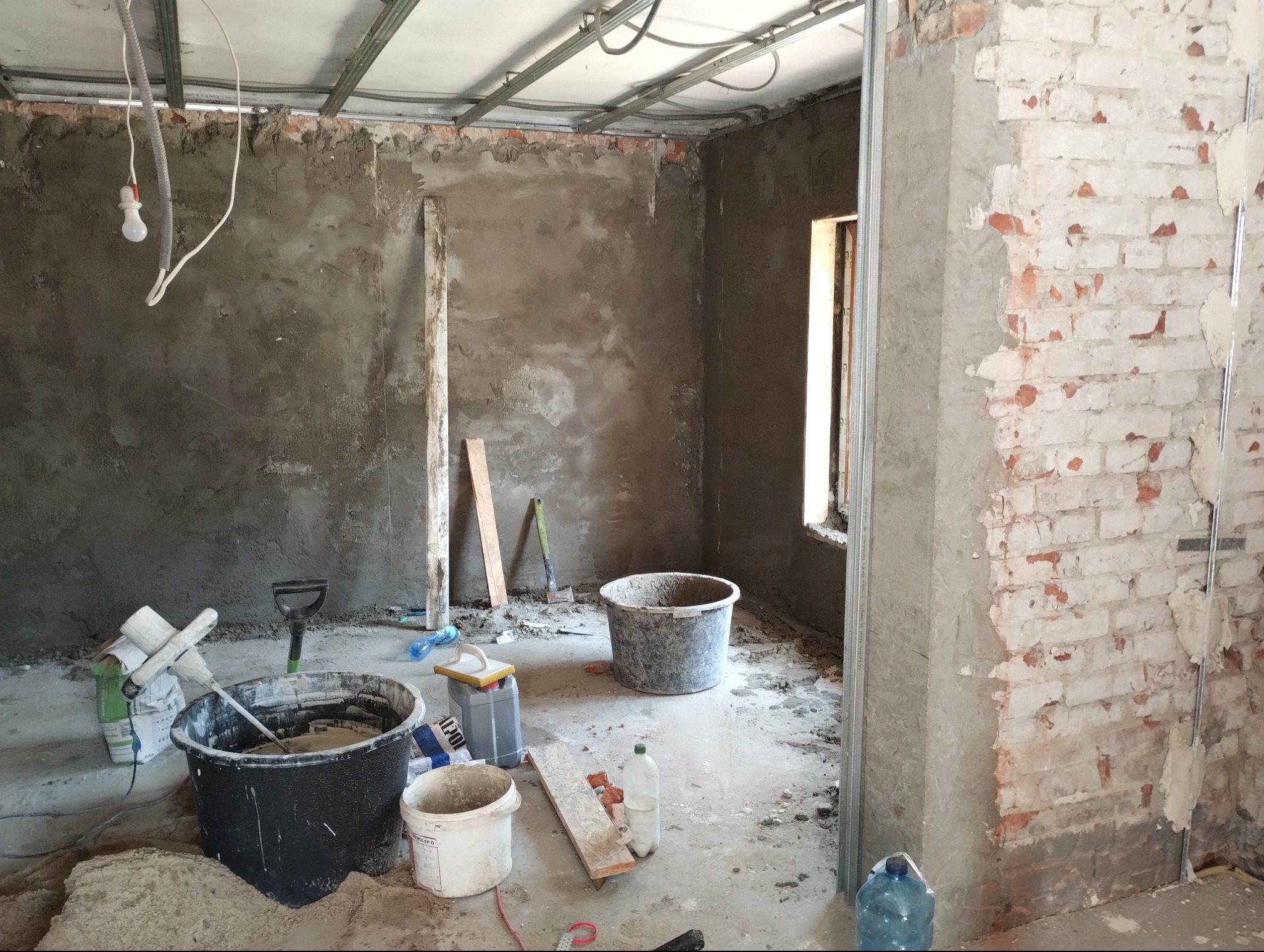 Wnętrze w trakcie prac przygotowawczych do wykończenia, widoczne surowe ściany, częściowo otynkowane, z odsłoniętą cegłą, konstrukcja sufitu podwieszanego, wiadra z zaprawą, mieszadło i narzędzia...