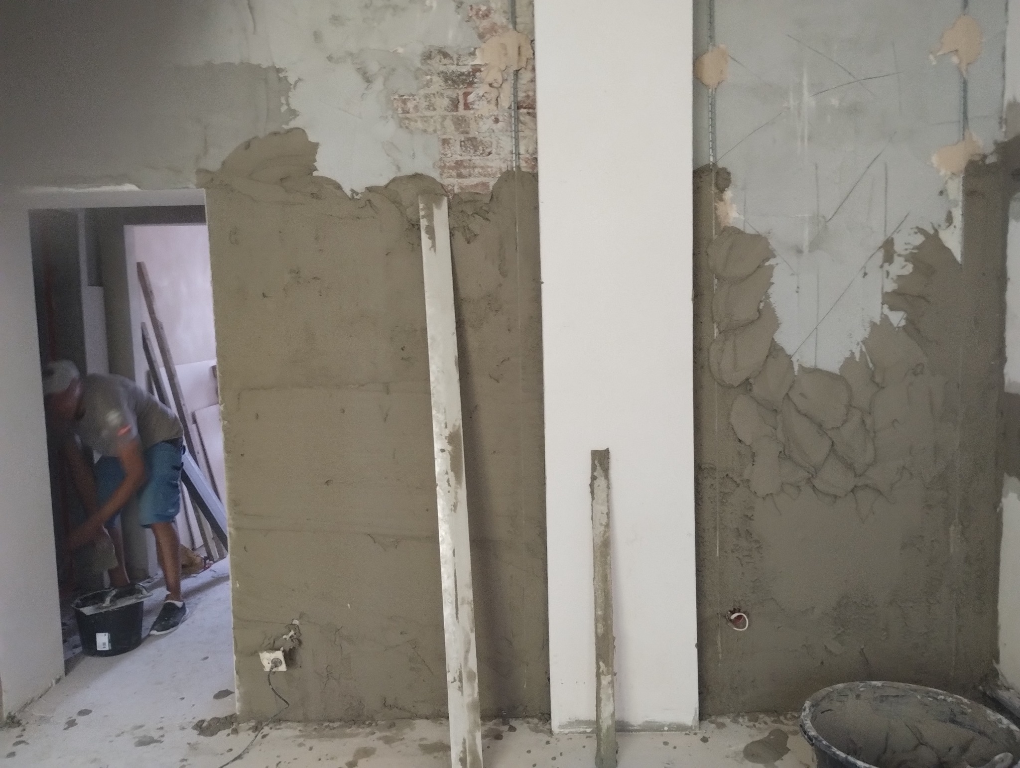 Przygotowanie ścian do położenia gładzi, widoczne profile pionowe, fragment odsłoniętej cegły i pracownik w tle mieszający zaprawę w wiadrze.