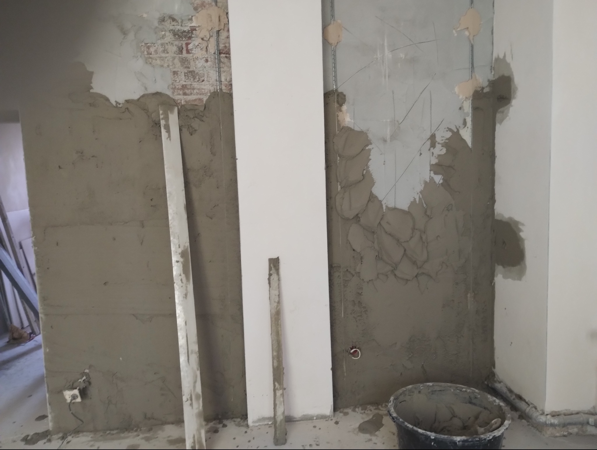Wnętrze w trakcie prac wykończeniowych: częściowo otynkowana ściana z fragmentami odsłoniętej cegły, gładka płyta oraz wiadro z zaprawą na podłodze.