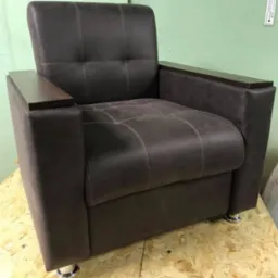 Brązowy fotel z drewnianymi podłokietnikami i chromowanymi nóżkami, widoczny na tle zielonej ściany i płyty OSB.