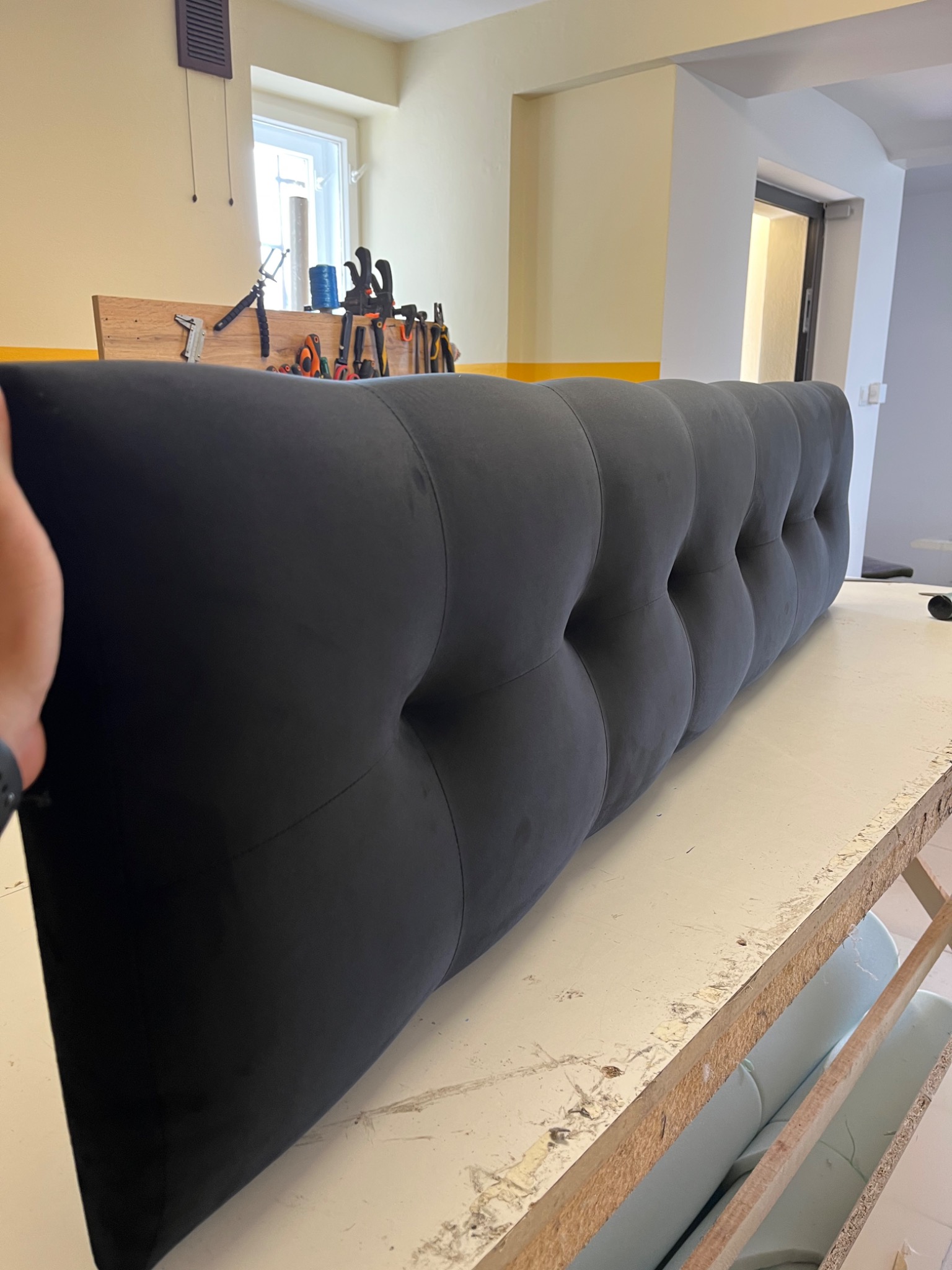 Gotowy, ciemnografitowy zagłówek łóżka z głębokimi pikowaniami, ustawiony na stole roboczym w warsztacie, w tle widoczne narzędzia i okno.